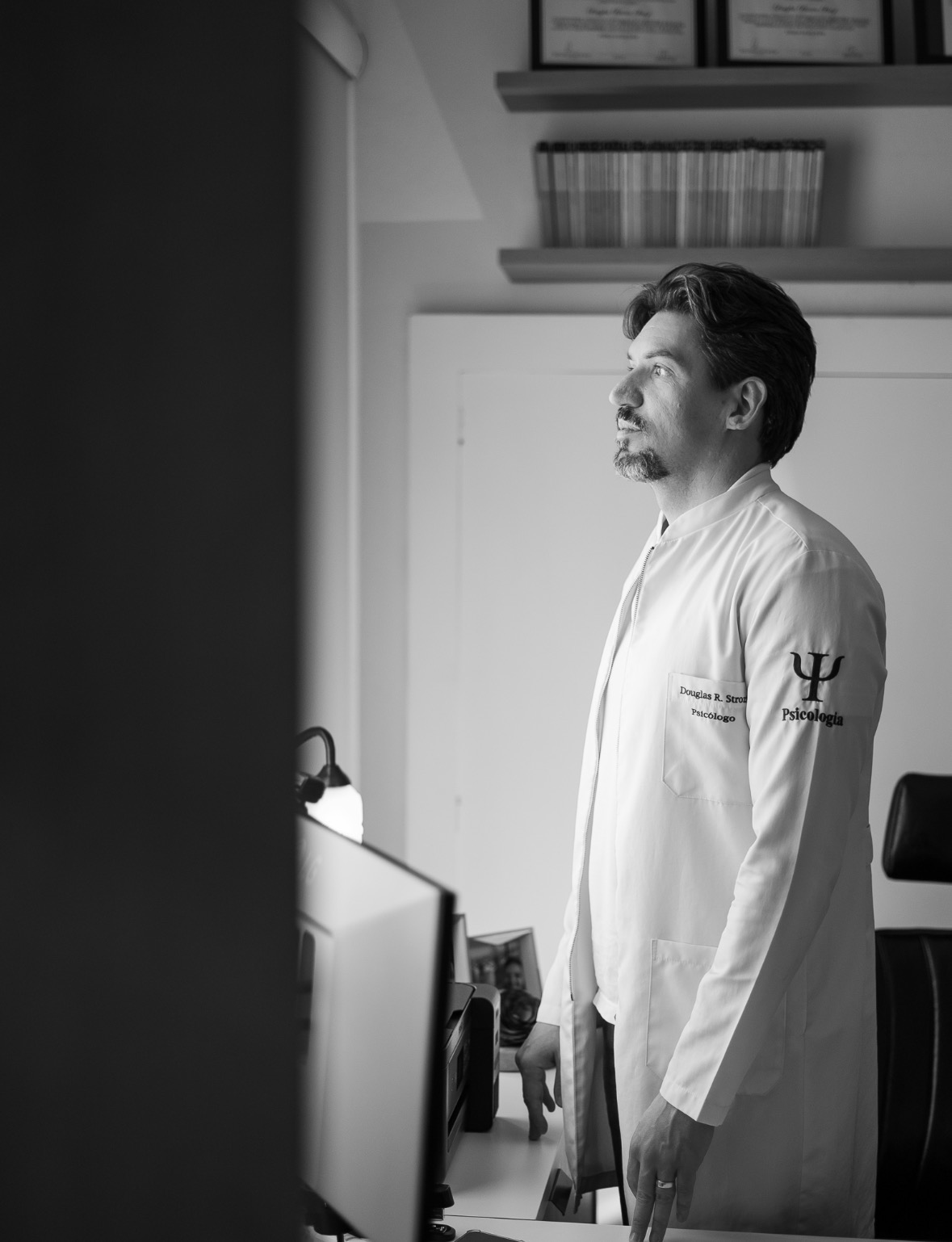 Douglas Renato Strozi de jaleco, em pé na porta do consultório, em fotografia em preto e branco